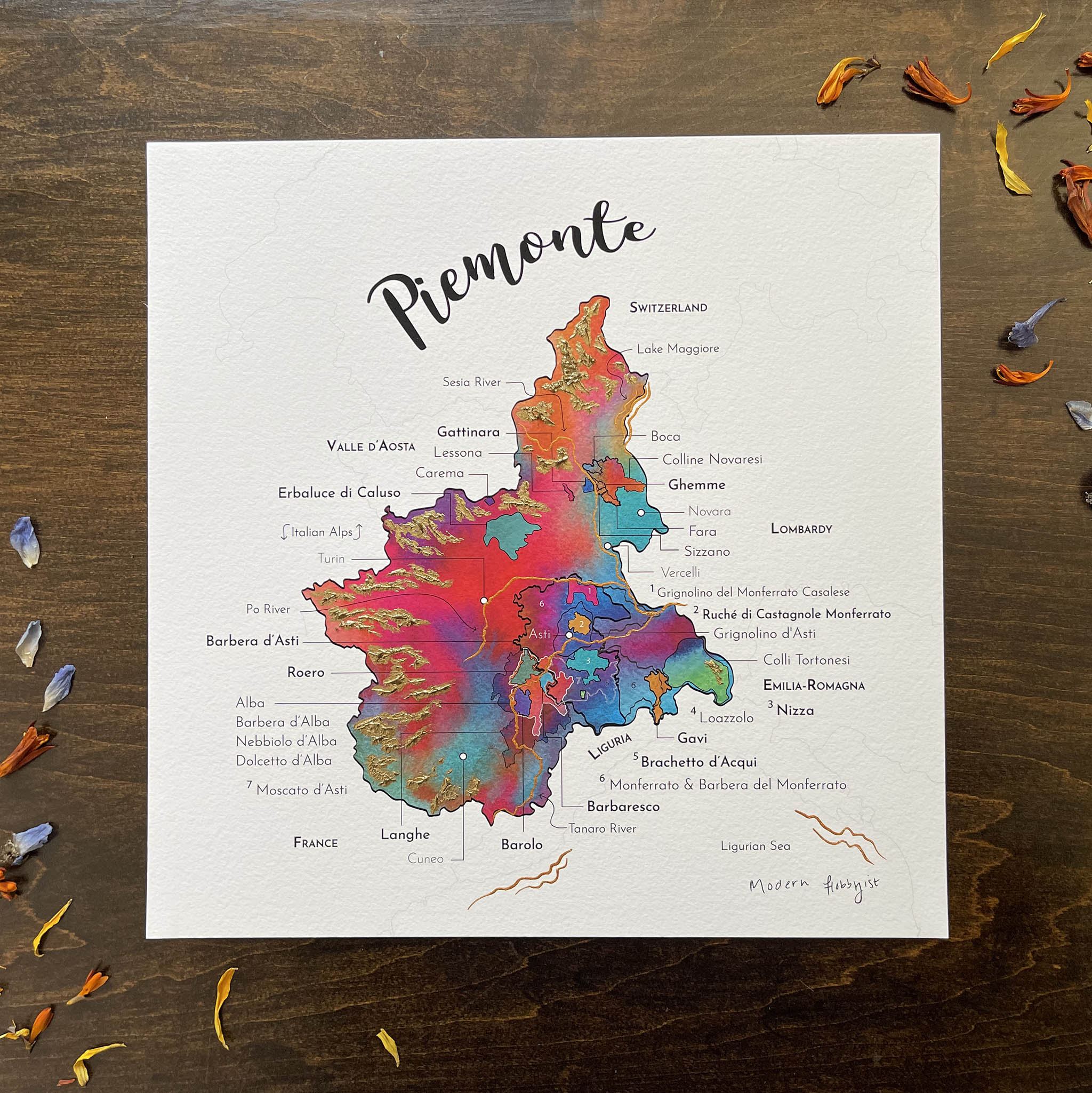 A colorful artistic representation of a map titled 'Piemonte' labeled with wine regions on a wooden surface surrounded by dried flower petals.